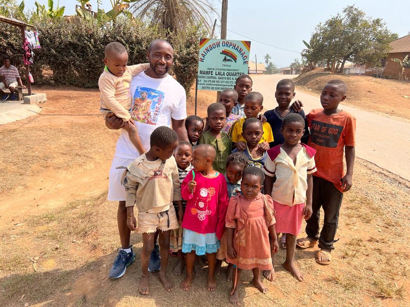 Kevin Collins visiting Rainbow Orphanage in Mamfe with children in front of the orphanage sign
